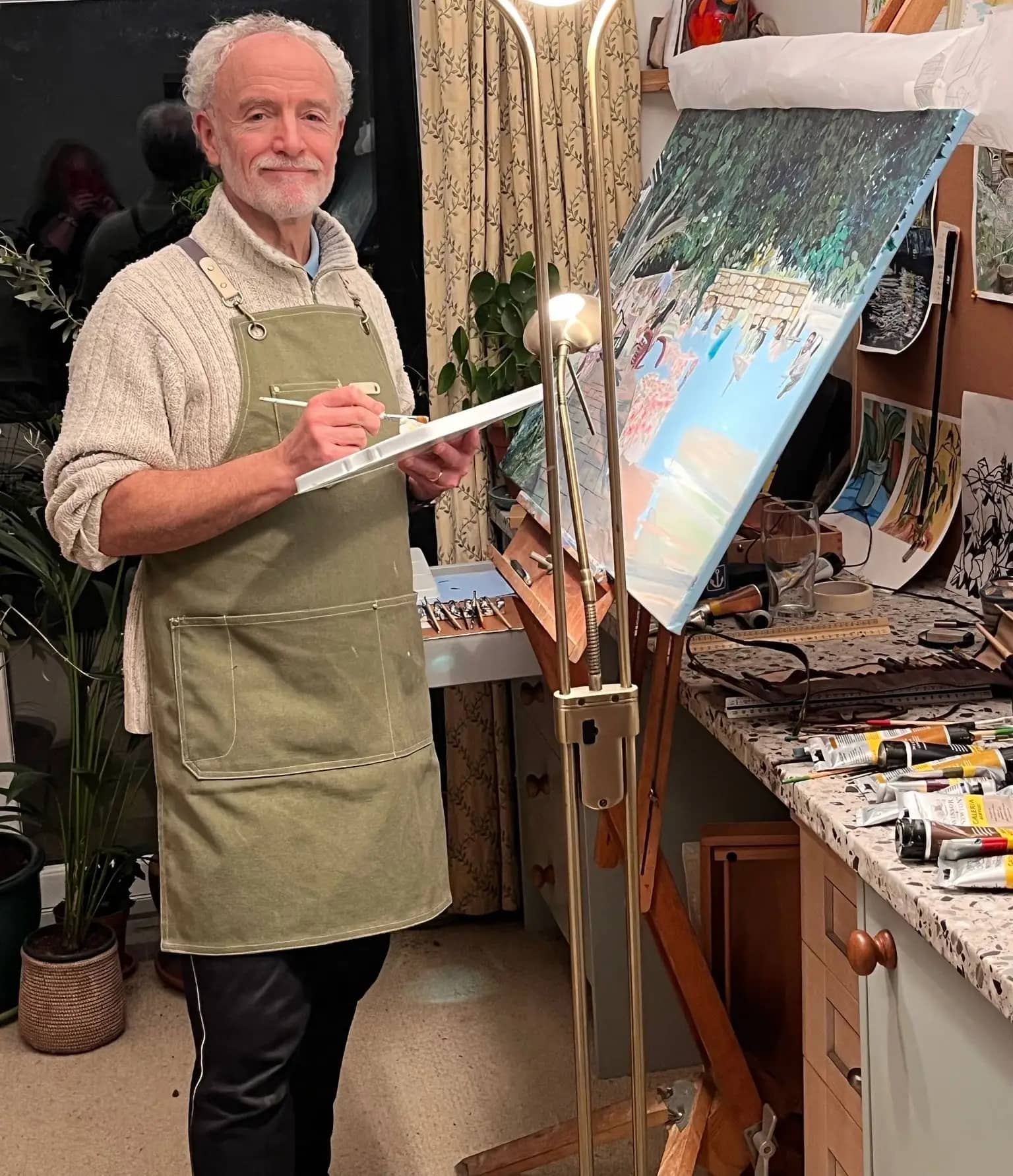 Peter Broad, a white man in his 60s with a crown of white hair, stands in front of an in-progress painting on an easel. He smiles gently towards the camera, paintbrush in hand.